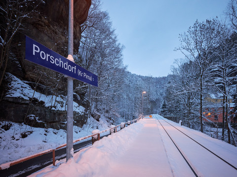 Haltestelle Nationalparkbahn  Verschneiter Bahnsteig in Porschdorf bei Dämmerung, Schild "Porschdorf" sichtbar.