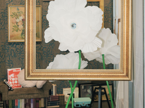 KSC_GUN_Best of II_Yablonovska_The Room_2025_FotoMarkFrost.jpg Weiße Blumen in einem Rahmen vor einer nostalgisch dekorierten Wand mit Gemälden und einem Sofa.White flowers in a frame in front of a nostalgically decorated wall with paintings and a sofa.Bílé květiny v rámu před nostalgicky vyzdobenou stěnou s obrazy a pohovkou.Białe kwiaty w ramce przed nostalgicznie udekorowaną ścianą z obrazami i sofą.Witte bloemen in een lijst voor een nostalgisch gedecoreerde muur met schilderijen en een bank.Fiori bianchi in una cornice davanti a una parete decorata in modo nostalgico con quadri e un divano.