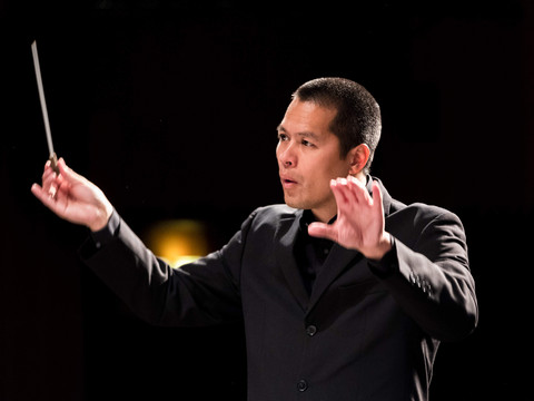 Portrait Chadik Ein Dirigent in formeller Kleidung mit erhobenen Händen und einem Taktstock beim Dirigieren.