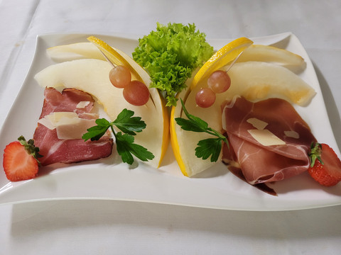 20230820_111623.jpg Das rechteckige Servierteller zeigt eine kunstvolle Anordnung von Schinken und Melonen, garniert mit grünem Salat, Erdbeerscheiben und Weintrauben.