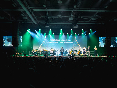 Oberberg singt Mitschnitt eines Konzerts mit Chor, Orchester und Lichtshow in vollem Veranstaltungsraum.