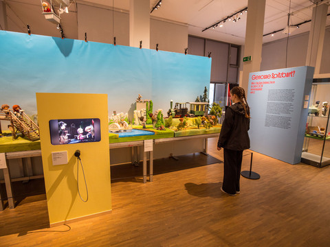 2 - Sandmännchen in Leipzig - Foto Andreas Schmidt.jpg Ein Ausstellungsraum mit Sandmännchen-Puppen und Kulissen zur Schau. Eine Frau betrachtet die Szene.An exhibition room with Sandman dolls and backdrops on display. A woman watches the scene.Výstavní místnost s vystavenými loutkami a kulisami Sandmana. Scénu sleduje žena.Sala wystawowa z lalkami i tłem Sandmana na wystawie. Kobieta przygląda się scenie.Een tentoonstellingsruimte met Sandman poppen en achtergronden. Een vrouw bekijkt het tafereel.Una sala espositiva con pupazzi e fondali di Sandman in mostra. Una donna osserva la scena.