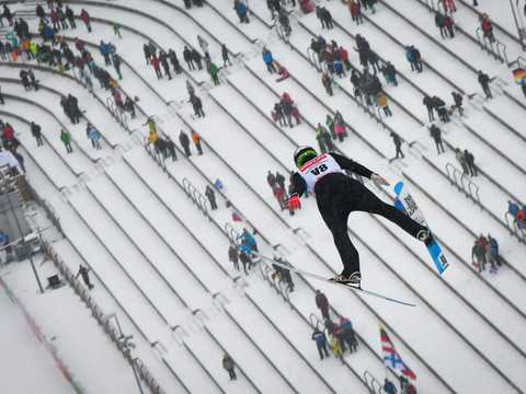 Sparkassen Vogtland Arena (c) Jan Simon Schäfer (1).jpg Ein Skispringer fliegt über die schneebedeckte Zuschauertribüne einer Sprungschanze.