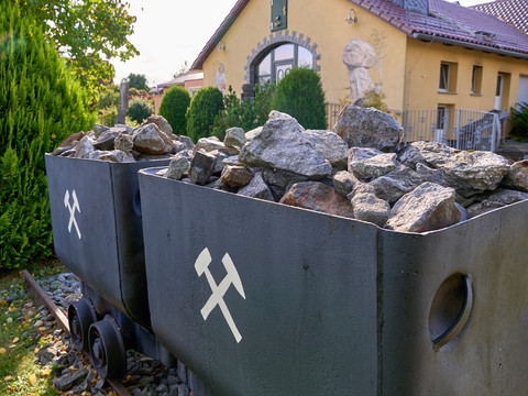 Gold-und-Mineralien-Erlebnisstaette-Neustadt-Yvonne-Brueckner-CCBY (2)-web.jpg Bergbauwagen Steinen und weißen Kreuzen als Zeichen vor einem Haus und Büschen.