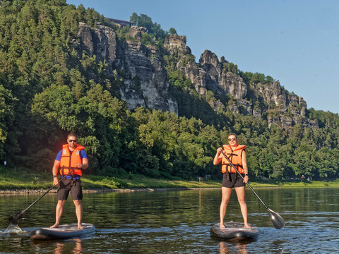 SUP_Elbe_(c)-TVSSW-Sebastian_Thiel_CC-BY-S2-TPR00786.jpg Zwei Personen auf Stehpaddelbrettern auf einem Fluss, bewaldete Klippen im Hintergrund.