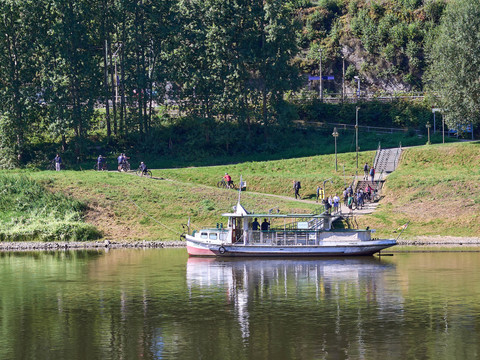 Boot mit Personen auf Fluss, Menschen am Ufer und Bäume im Hintergrund.