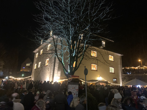 Bielsteiner Weihnachtsmarkt Blick auf ein mit Schweinwerfern angestrahltes Haus. davor steht ein Baum und es sind zahlreiche Menschen zu sehen, die sich um das Haus herum aufgestellt haben.
