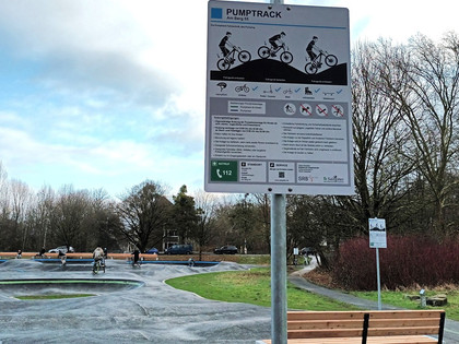 Die Pumptrackanlage in Salzgitter-Gebhardshagen Infoschild und Holzbank an einer asphaltierten Pumptrackanlage mit Radfahrern.