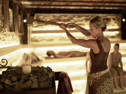 Mediterana Aufguss Eine Frau steht in einer Sauna und mach einen Aufguss. Im Hintergrund sind noch drei weitere Personen zu erkennen.