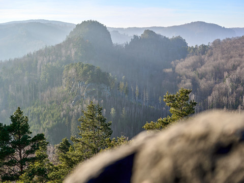Blick auf bewaldete Hügel und Felsen, Vordergrund unscharf, unter blauem Himmel.