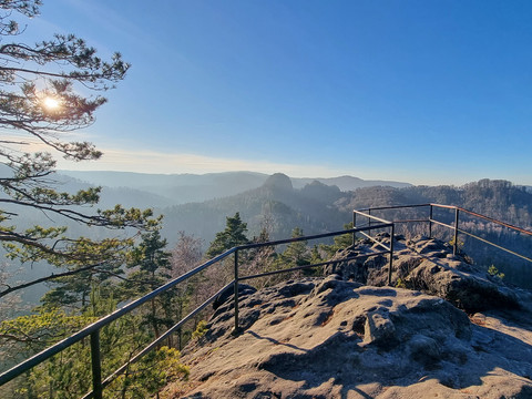 Aussichtspunkt auf einem Felsen mit Geländer, Blick auf bewaldete Hügel im Sonnenlicht.
