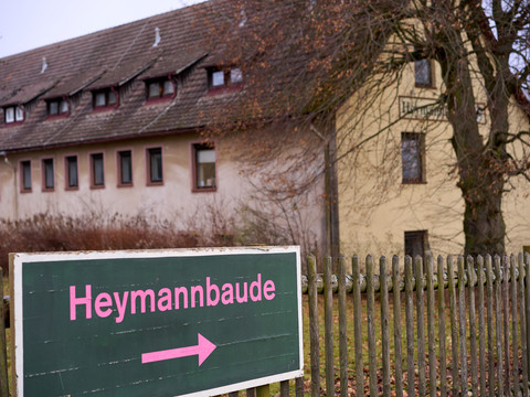 Wegweiser "Heymannbaude" mit Pfeil nach rechts, dahinter gelbes Gebäude und Zaun.