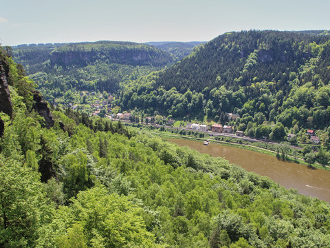 Blick auf ein grünes Tal mit Fluss, umgeben von bewaldeten Hügeln und Felsen.