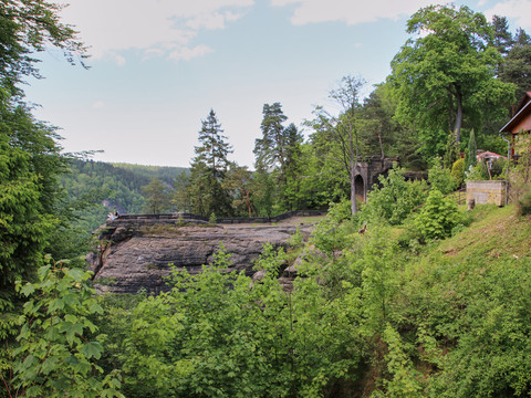 Waldlandschaft mit Holzhaus und Felsvorsprung, im Hintergrund eine Aussichtspattform.
