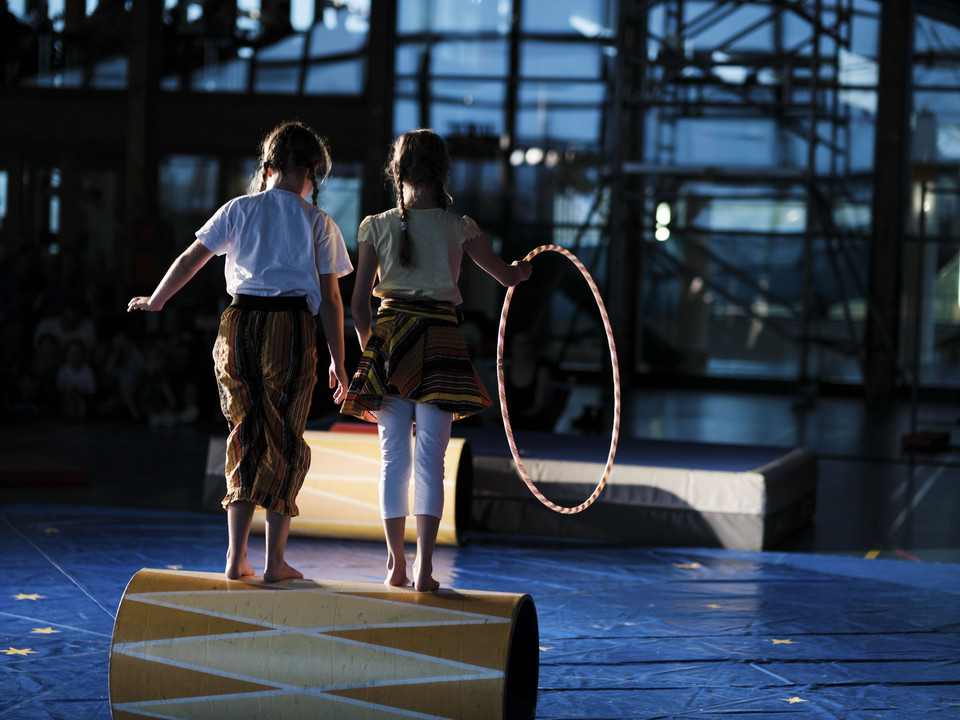 Gemeinsam auf der Lauftrommel Zwei Mädchen balancieren gemeinsam auf einer gelben Lauftrommel im Mitmachzirkus Aletsch Arena vor grossen Fenstern.Two girls balance together on a yellow walking drum in the Aletsch Arena hands-on circus in front of large windows.Deux filles se tiennent en équilibre sur un tambour de course jaune dans le cirque participatif Aletsch Arena devant de grandes fenêtres.