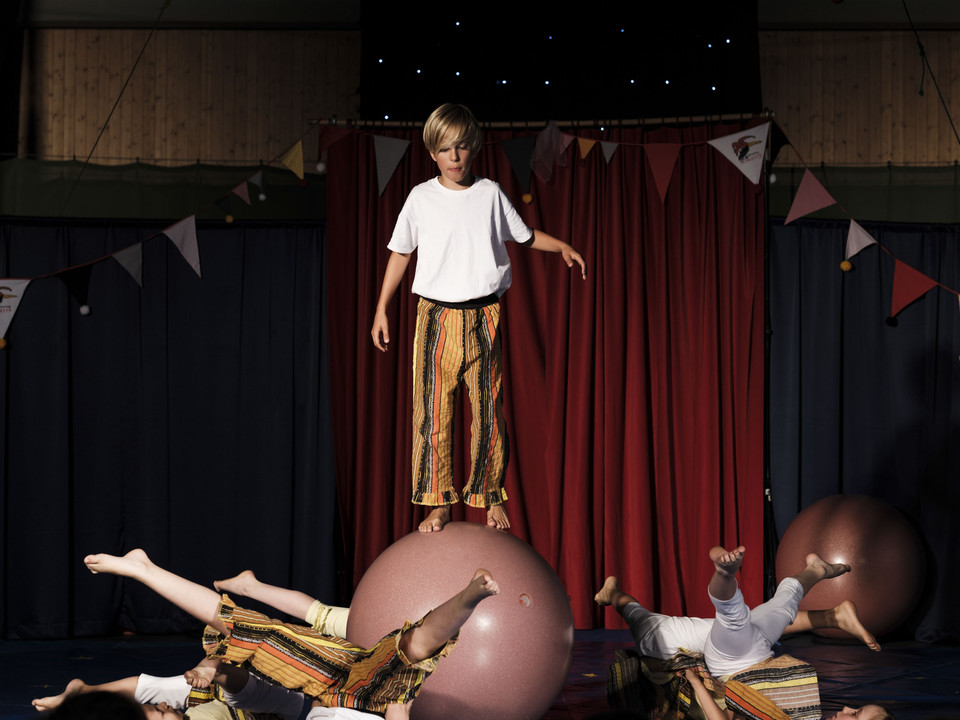 Gruppenakrobatik in der Manege Ein Junge balanciert auf einer Kugel, umringt von liegenden Kindern bei einer Show im Mitmachzirkus Aletsch Arena.A boy balances on a ball surrounded by reclining children during a show at the Aletsch Arena hands-on circus.Un garçon en équilibre sur une boule, entouré d'enfants allongés, lors d'un spectacle au cirque participatif Aletsch Arena.