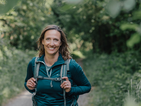 Naturcoach Christiane Kaiser Christiane Kaiser in Wanderkleidung und mit Rucksack auf einem bewaldeten Pfad.