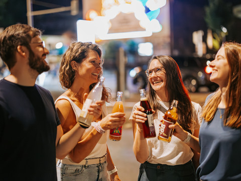 Nachtleben auf der KarLi - Szenemeilen in Leipzig Eine Gruppe Erwachsener steht abends mit Getränken in der Hand vor dem Leuchtschild "Löffelfamilie" an der Feinkost auf der Karl-Liebknecht-Straße