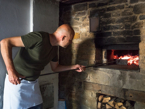 historisches Backhaus im LVR-Freilichtmuseum Lindlar Mann in grünem T-Shirt und weißer Schürze schürt Feuer in einem traditionellen Steinofen mit Glut.