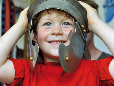 Ein lachender Junge trägt einen antiken Heldenhelm und blickt fröhlich in die Kamera.