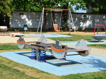 Landrattenspielplatz_Thiede (6)_HD_16zu9.JPG Spielplatz mit hölzernem Wippschiff auf blauen Fallschutzmatten, Schaukel und Sitzbank im Grünen.