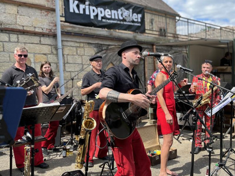 Band mit Musikern in roten Hosen, die auf einer Bühne spielen. Sänger mit Gitarre und Sängerin mit Mikrofon.