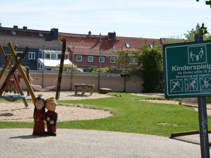 Roxy-Helco-Spielplatz1_HD_16zu9.JPG Spielplatz mit Holzgeräten, Sandflächen, Rasen, zwei Holzfiguren und Hinweisschild im Freien.