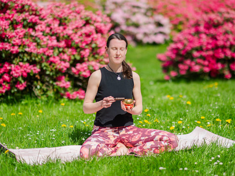 Meditation im Schneidersitz Frau sitzt im Schneidersitz auf einer Matte, meditiert mit einer Schale in der Hand, umgeben von blühenden Blumen.