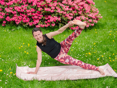 Seitstütz Frau in Yogahaltung auf einer Matte, umgeben von bunten Blumen und grünem Gras.