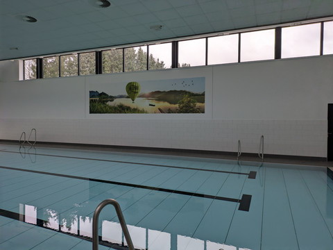 Innenbereich Hallenbad Hallenbad mit blauem Wasser, Bahnen, Treppen und Wandbild einer Seenlandschaft mit Heißluftballon.