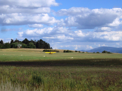 Flugplatz_Schaeferstuhl_Salzgitter_8_HD_16zu9.JPG Gelbes Sportflugzeug auf einer grünen Graspiste vor Bäumen und Bergen unter bewölktem Himmel.