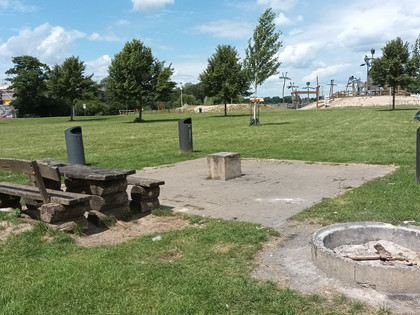 Grillplatz_Piratenspielplatz_HD_16zu9.jpg Grillplatz auf einer Wiese mit steinerner Feuerstelle, rustikalen Holzbänken und Mülleimern.