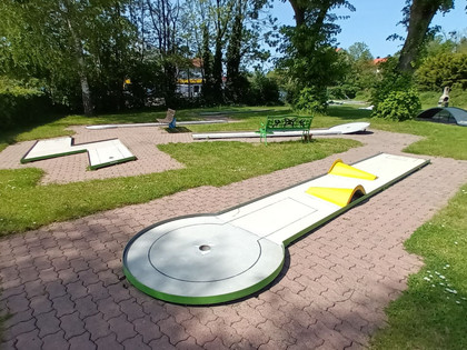 Minigolfanlage_Lebenstedt_HD_16zu9.jpg Mehrere helle Minigolfbahnen mit Hindernissen auf grünem Rasen zwischen Bäumen.