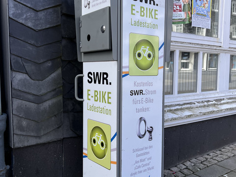 E-Bike-Ladestation am Marktplatz E-Bike-Ladestation am Marktplatz mit Infotafeln und Stromanschluss vor einem Schiefergebäude.