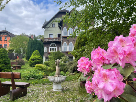 Margaretenhof Gohrisch Außenansicht Blühende rosa Rhododendren im Vordergrund, im Hintergrund ein Garten mit Bäumen und einem Haus.