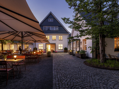 Gourmetrestaurant „Schote“ Beleuchteter Innenhof mit Tischen, großen Schirmen und historischem Gebäude bei Abenddämmerung