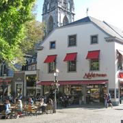 Nobis Printen Münsterplatz Auf dem Münsterplatz befindet sich das große Haus der Bäckerei Nobis mit vielen Sitzgelegenheiten davor, die zur Hälfte mit Kunden belegt sind. Links ragt ein blühender Baum ins Bild.