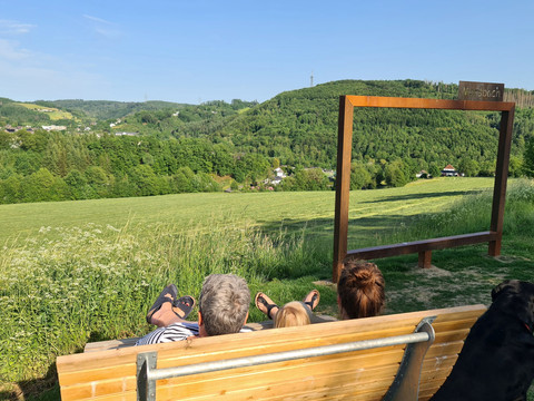Waldsofa und Fotorahmen Personen auf Holzbank blicken über grüne Hügellandschaft mit großem Metallrahmen im Vordergrund