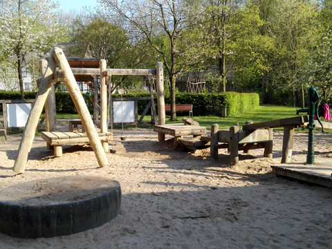 Kinderspielplatz Alte Dombach  Klettergerüst aus Holz und Balancierbrücke auf einem sandigen Spielplatz, umgeben von Bäumen.