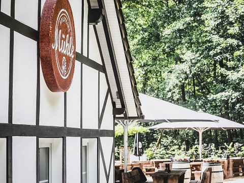 Historische Holstein's Mühle Fachwerkgebäude mit "Mühle" Schild, Terrasse und Sitzgelegenheiten in grüner Waldkulisse.