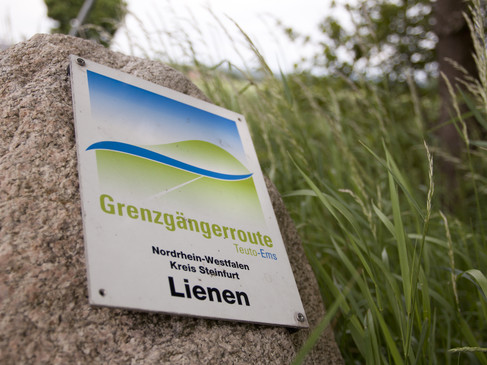 Schild der Grenzgängerroute Teuto-Ems in Lienen, umgeben von hohen Gräsern.