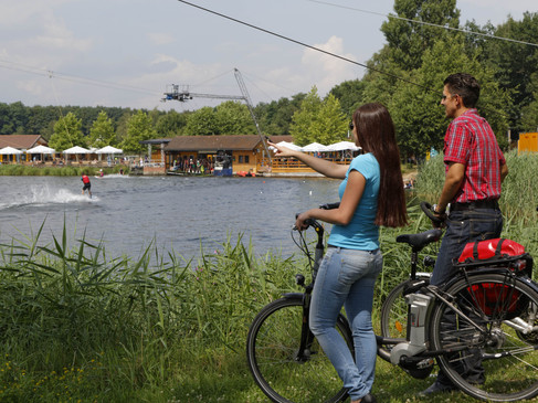 Zwei Personen beobachten eine Wakeboarder am See, im Hintergrund eine Seilbahn.