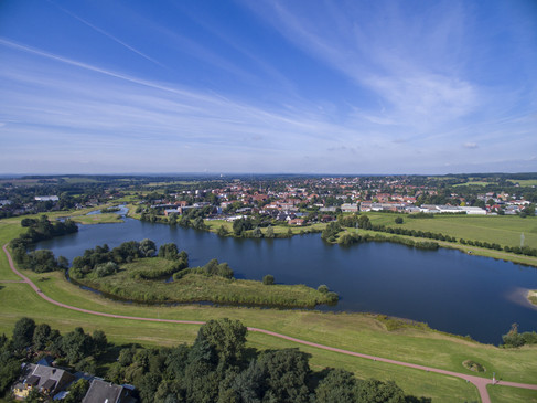 Luftaufnahme einer weitläufigen Landschaft mit See, Grünflächen und einer Stadt im Hintergrund.