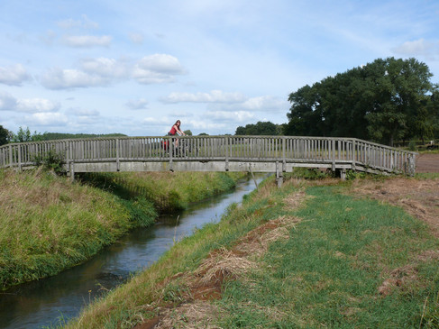 Holzbrücke über einen kleinen Fluss, flankiert von grünen Wiesen unter einem wolkigen Himmel.Wooden bridge over a small river, flanked by green meadows under a cloudy sky.Træbro over en lille flod, flankeret af grønne enge under en overskyet himmel.Houten brug over een kleine rivier, geflankeerd door groene weiden onder een bewolkte hemel.