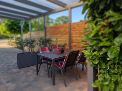 2 Terrasse 2.png Überdachte Terrasse mit Tisch, vier Stühlen, Pflanzen und Sichtschutz aus Holz.