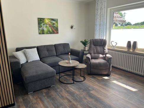3 Wohnzimmer 3.jpg Wohnzimmer mit grauem Ecksofa, Sessel, runden Tischen und großem Fenster.