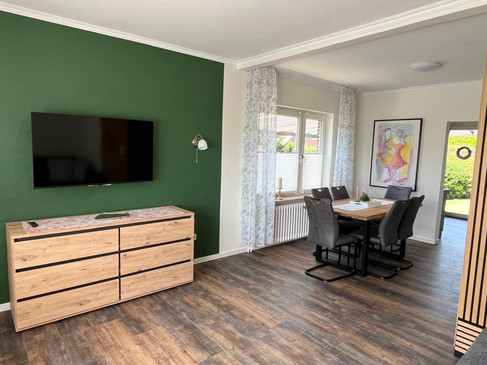 4 Essecke .jpg Wohn- und Essbereich mit Holztisch, grauen Stühlen, TV und grüner Akzentwand.