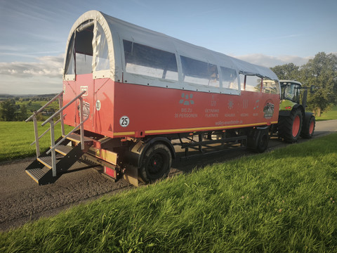 Wolle's Eventfahrten Roter Planwagen hinter Traktor auf Landstraße in grüner Hügellandschaft