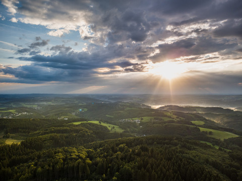 Reichshof Blockhaus Sonnenstrahlen durchbrechen dramatische Wolken über einer weitläufigen, grünen Hügellandschaft.