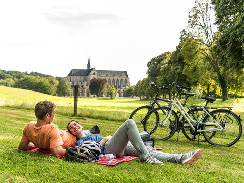 Rad fahren in Altenberg Paarsitzung auf grüner Wiese mit Blick auf historische Abtei; zwei Fahrräder stehen daneben.